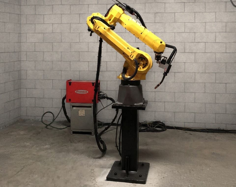 Six-axis robotic welding arm on a steel pedestal in a workshop.